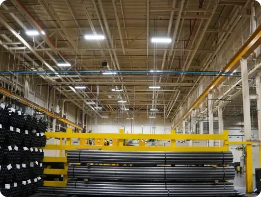 Warehouse interior with stacked metal rods and beams. Overhead lights illuminate the spacious building.