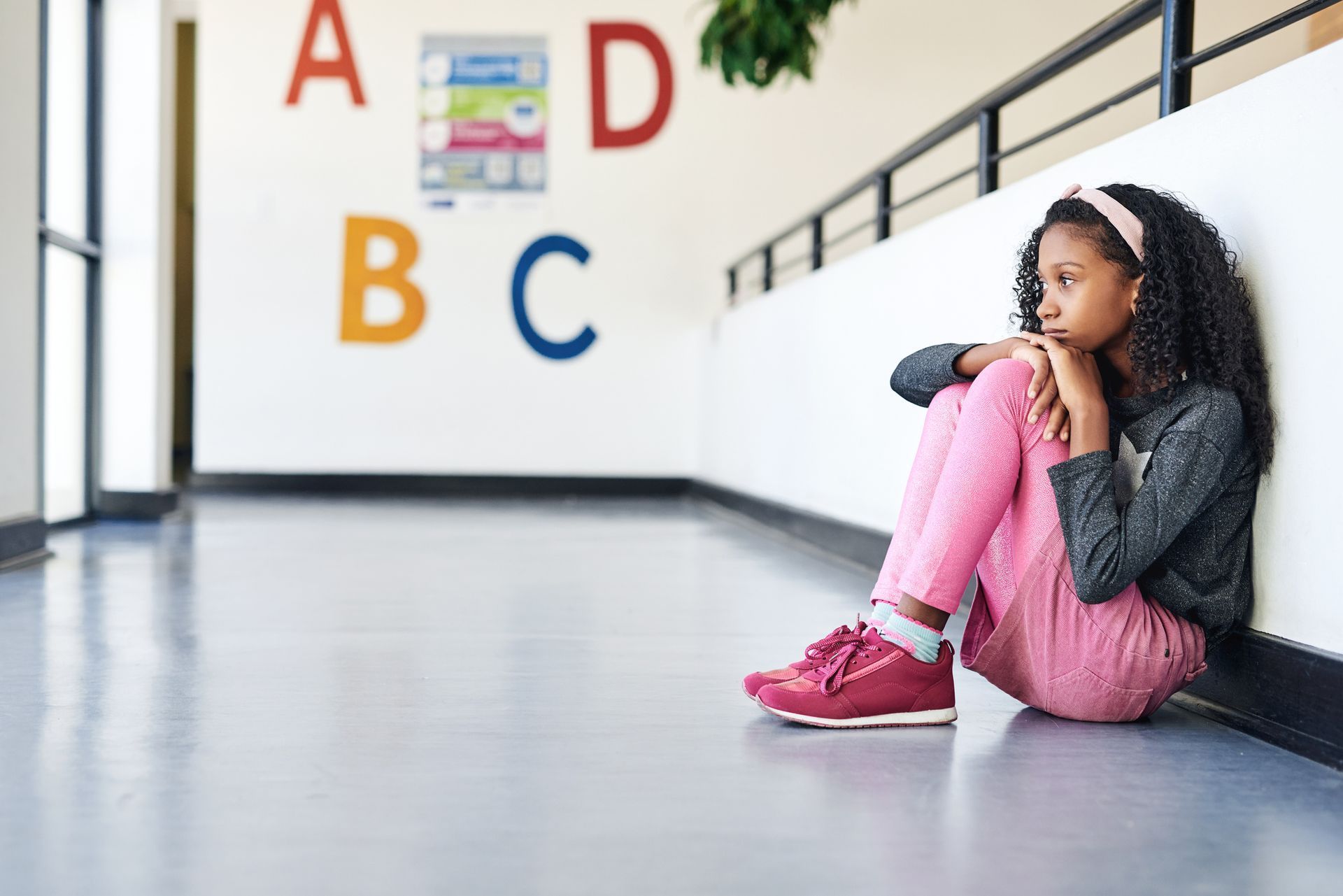 A young girl is sitting on the floor leaning against a wall in a hallway.