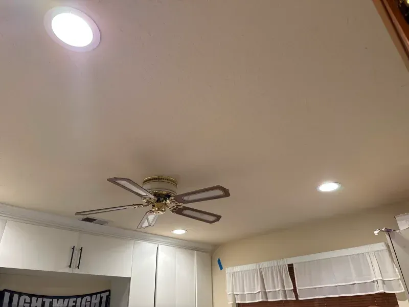 A ceiling with recessed lighting and a ceiling fan, above white cabinets and a window with curtains.