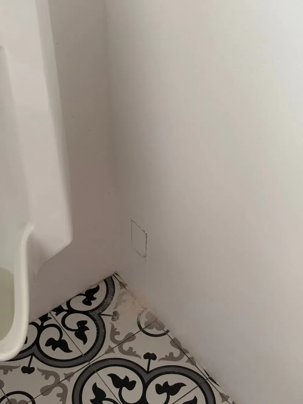 White wall corner with access panel, patterned floor tiles, and a white fixture.