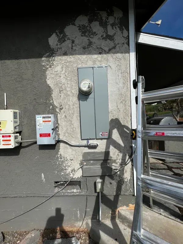 Electrical panel and meter on a stucco wall. Gray panel box and meter, damage around it. Ladder leans against the building.