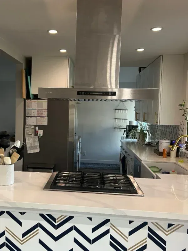 Kitchen with stainless steel range hood, cooktop, and chevron-patterned countertop. Refrigerator and sink are visible.