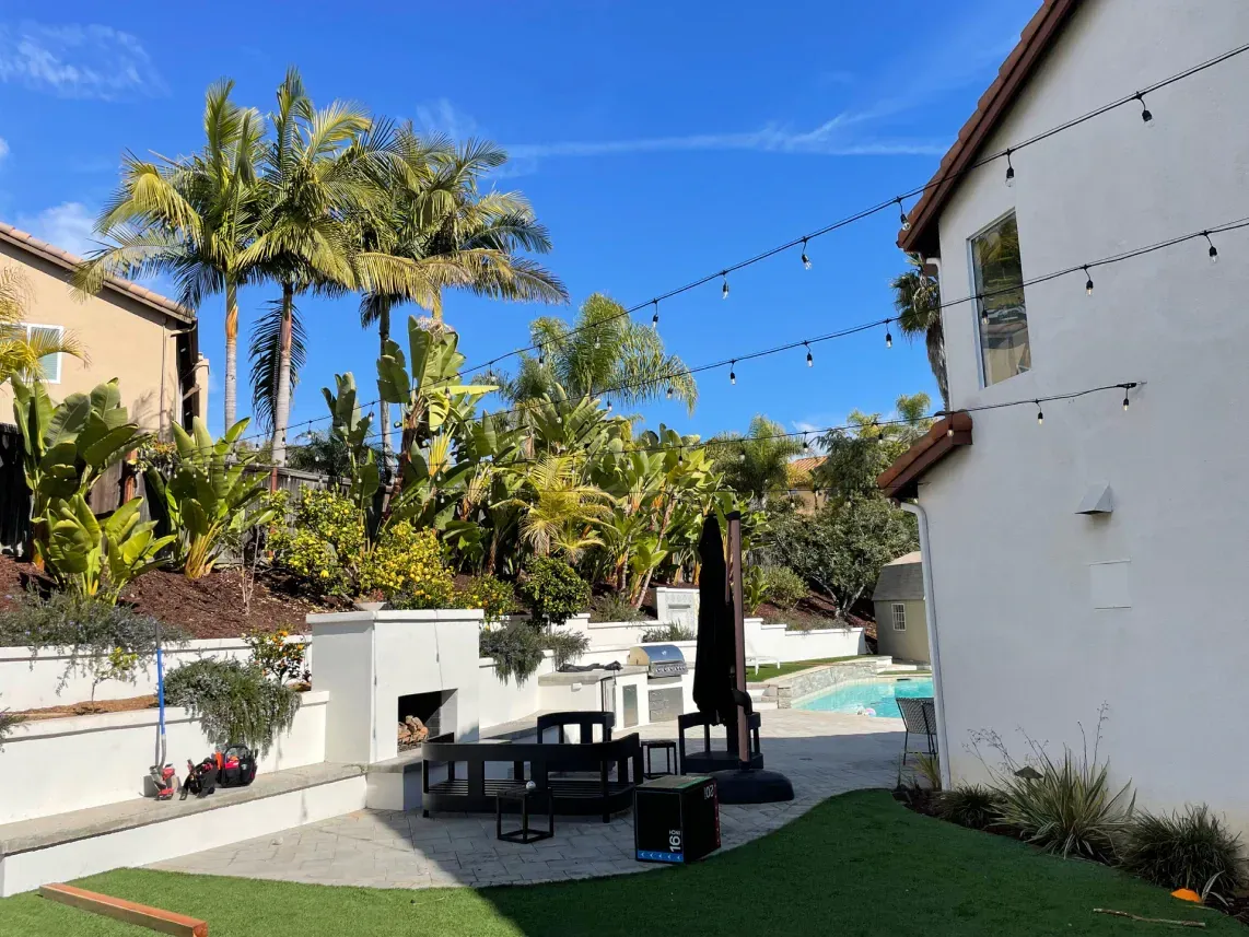 Backyard patio with string lights, palm trees, outdoor kitchen, seating, and pool under blue sky.
