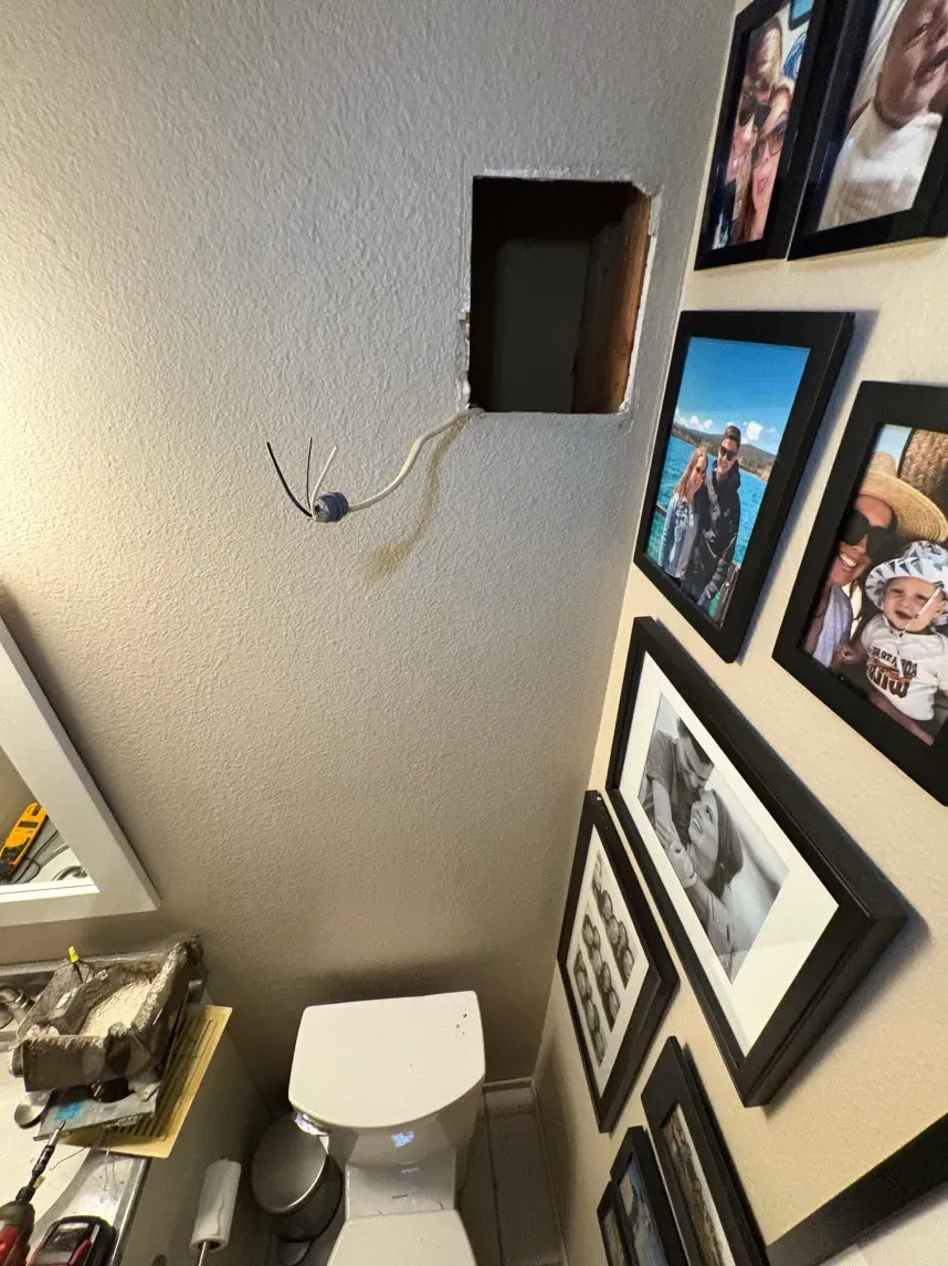 Hole in wall above toilet with exposed wires.  Photo frames on adjacent wall.  Bathroom setting.