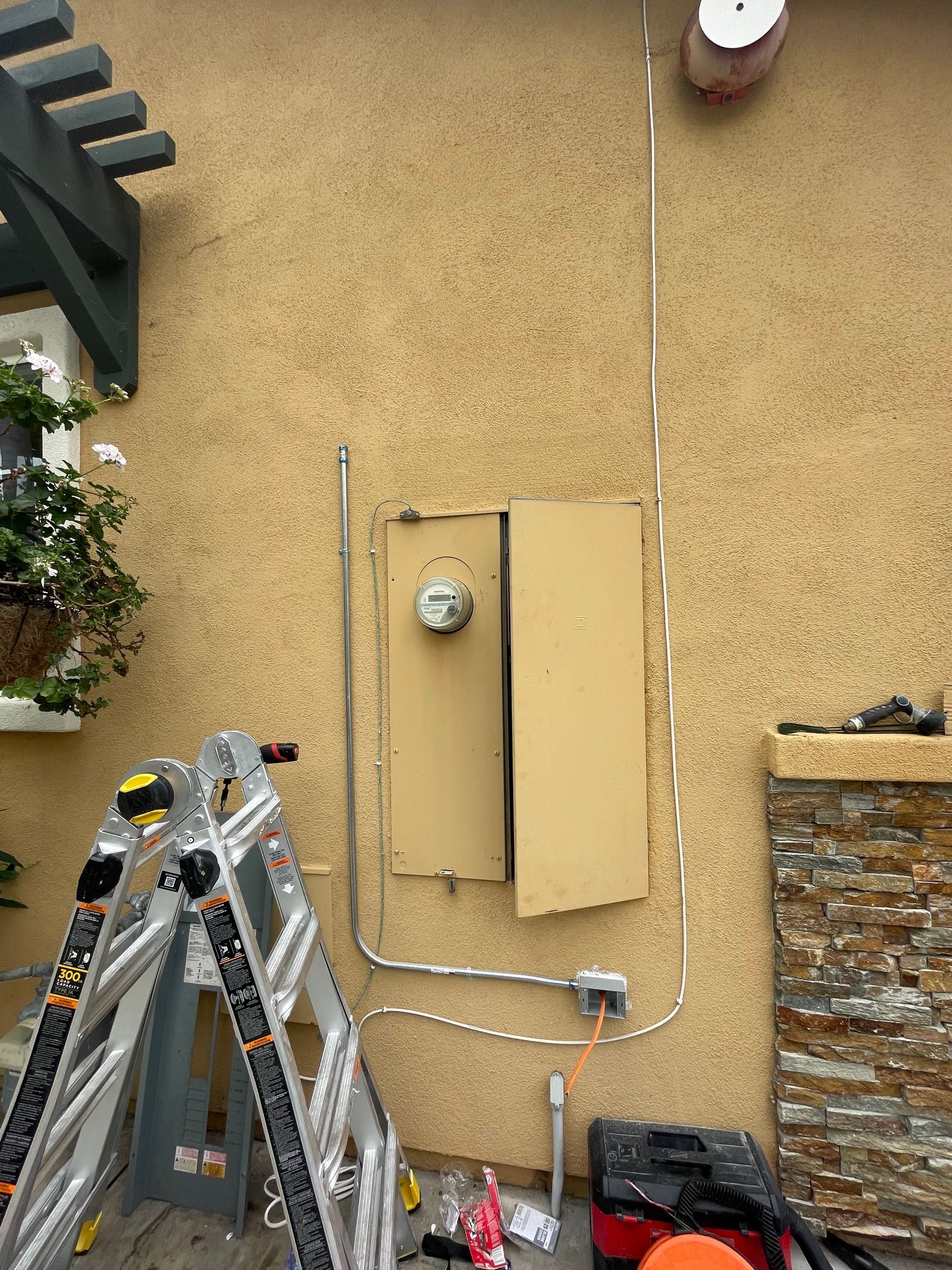 Electrical meter and panel on a tan stucco wall, with a ladder and tools below.