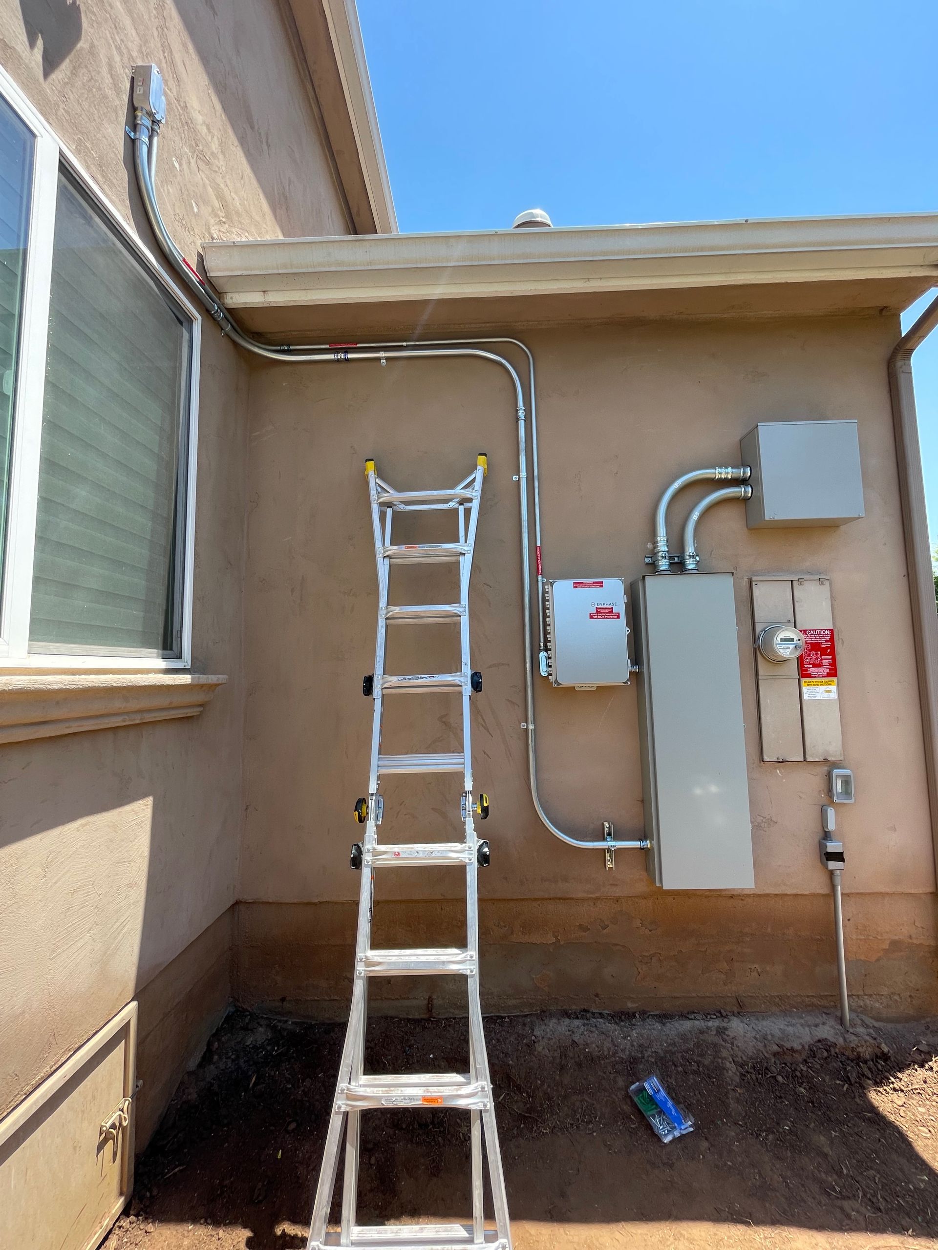 Exterior wall with electrical equipment, conduit, and ladder. Sunny day, beige stucco.