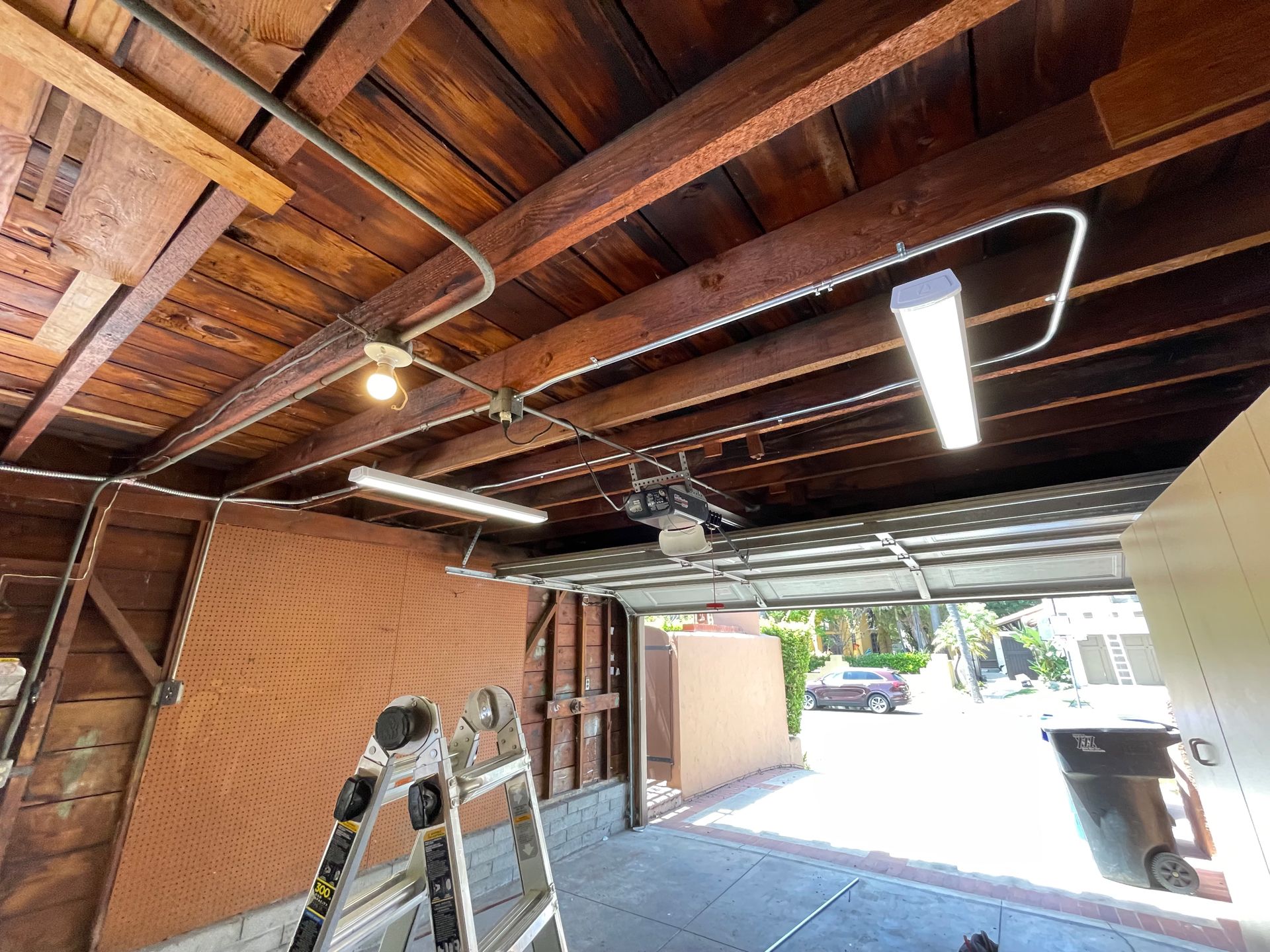 Garage interior with exposed wooden beams, fluorescent lights, and open garage door.