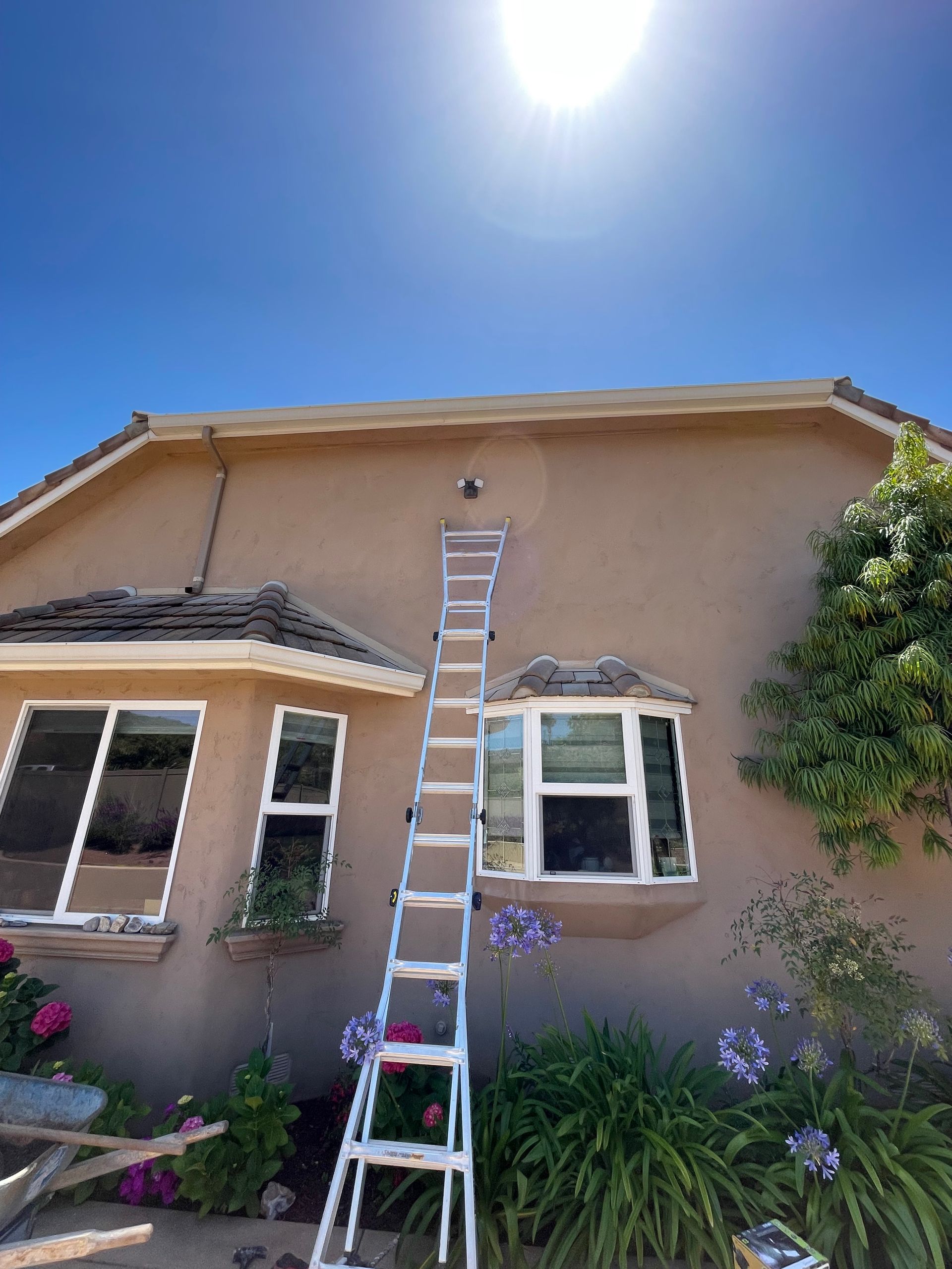 Ladder against a beige house with a clear blue sky, reaching towards the roofline.