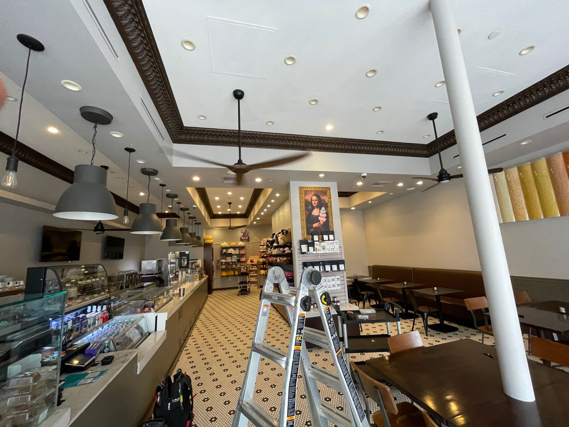 Interior of a cafe with a ladder, display cases, tables, and ceiling lights.