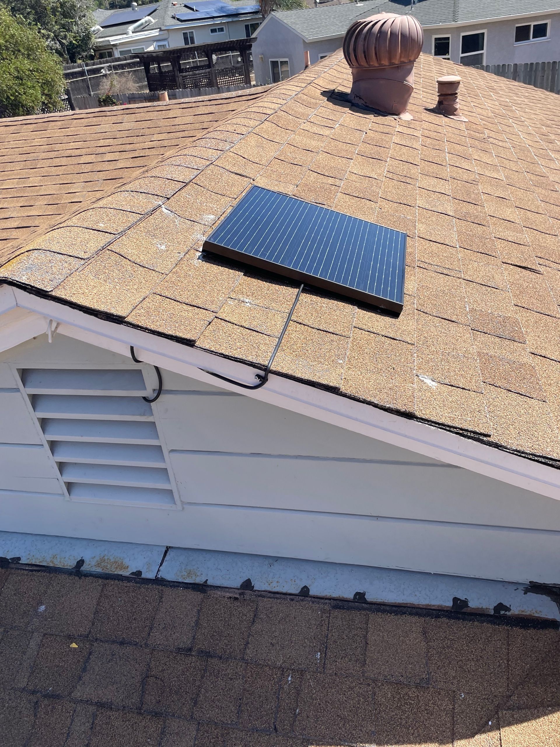 Solar panel on a brown shingled roof next to a vent, a white building.