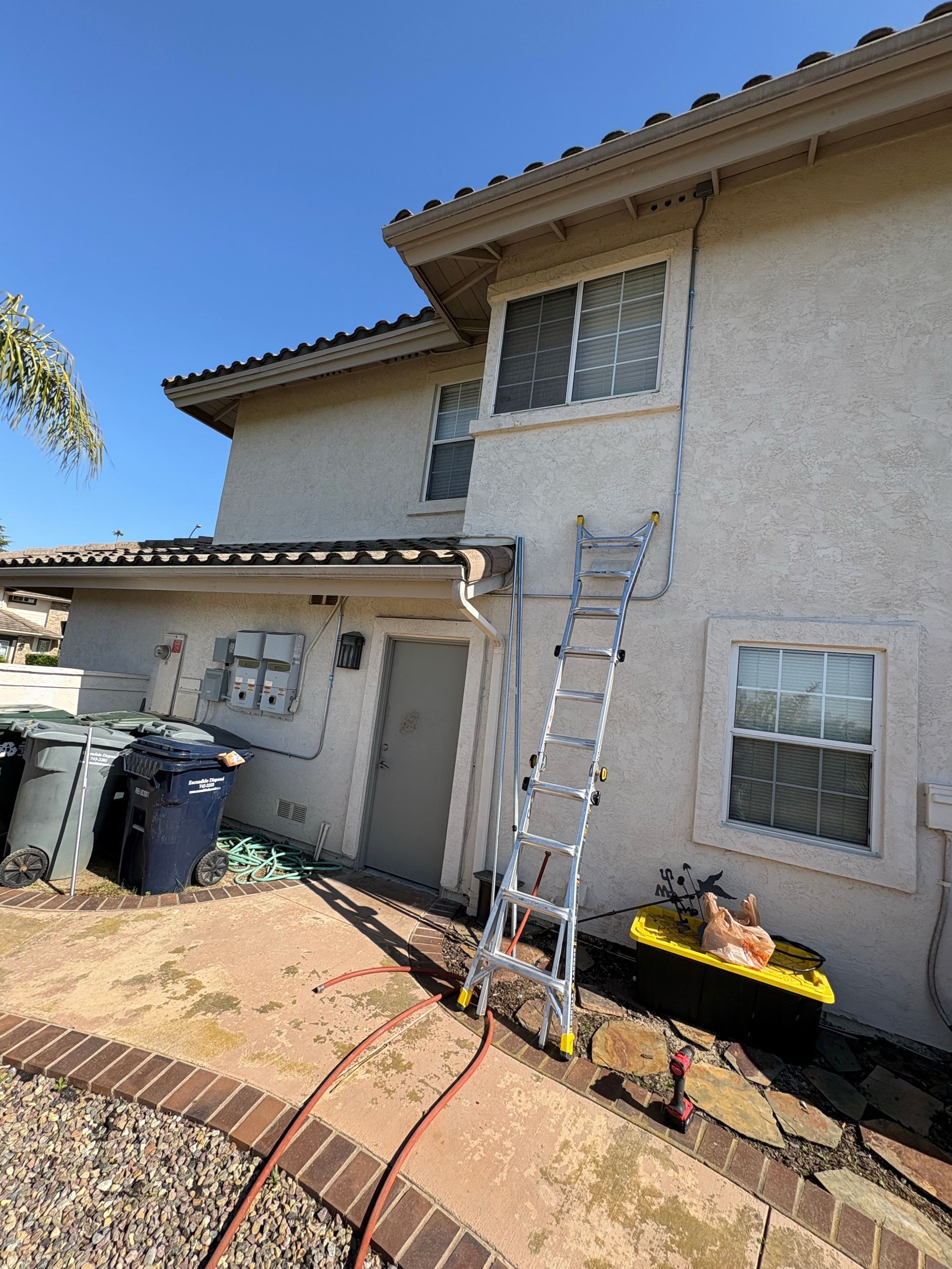 Ladder leaning against a two-story building with stucco walls, near trash bins, and a hose on the ground.