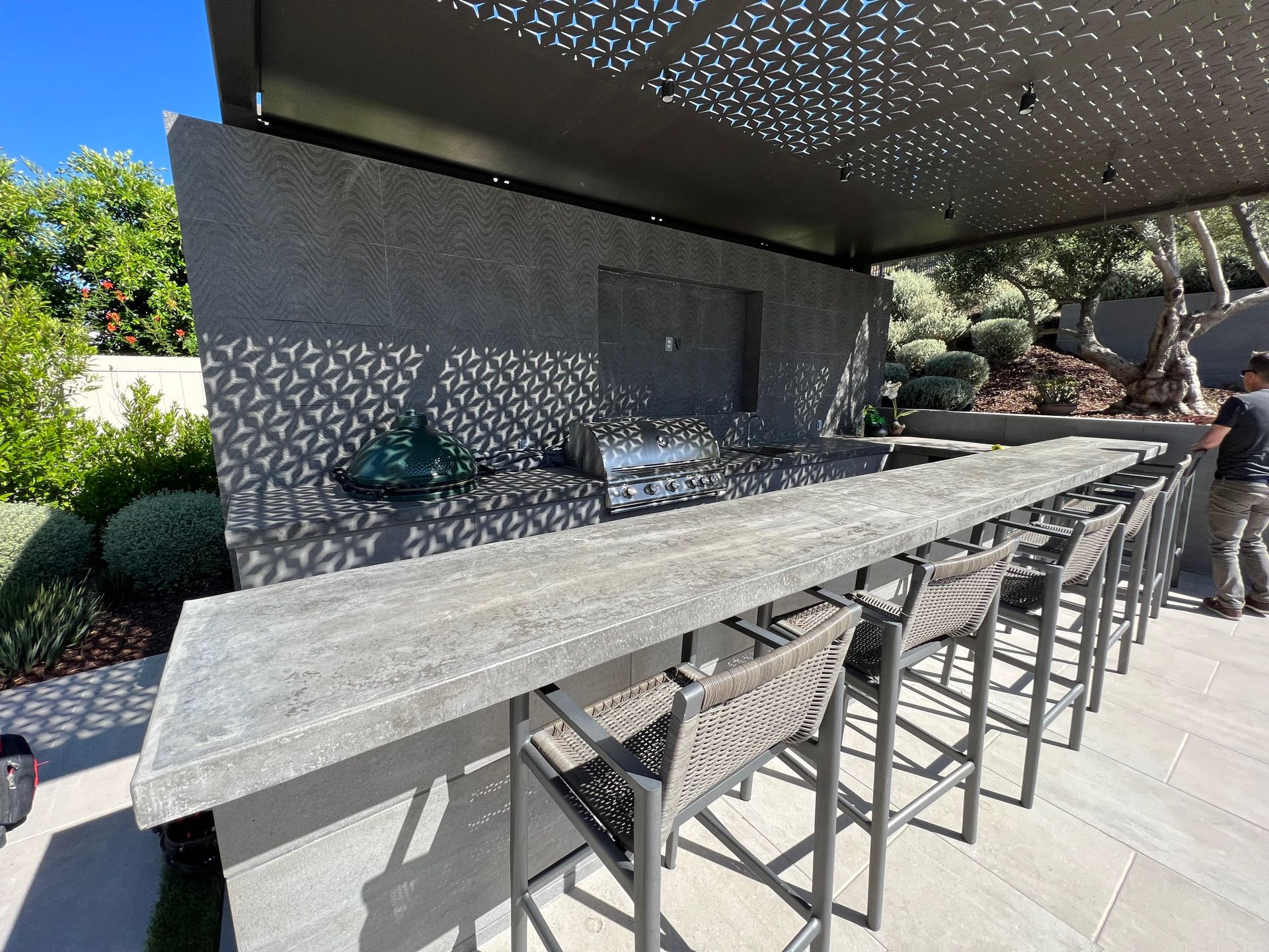 Outdoor bar with concrete countertop, metal stools, and decorative overhead canopy.
