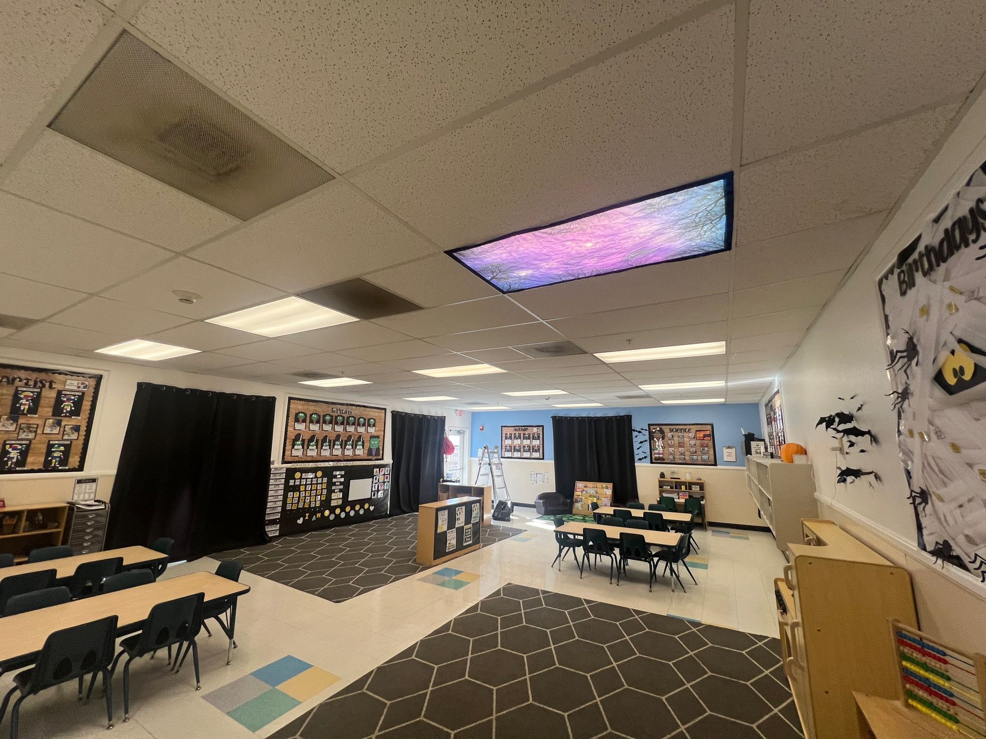 Classroom interior, black curtains, tables, and ceiling lights, with patterned carpet and posters on the walls.