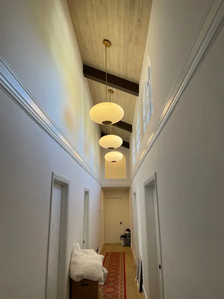 Hallway with white walls, wood ceiling, pendant lights, and doors. A person sits at the end.