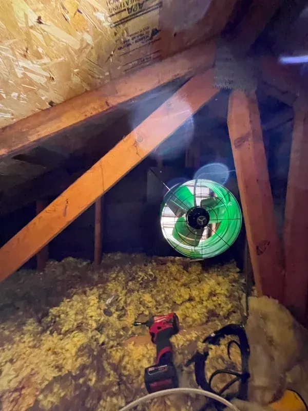 Attic with wooden beams, insulation, and a green-lit exhaust fan. A drill lies on the insulation.