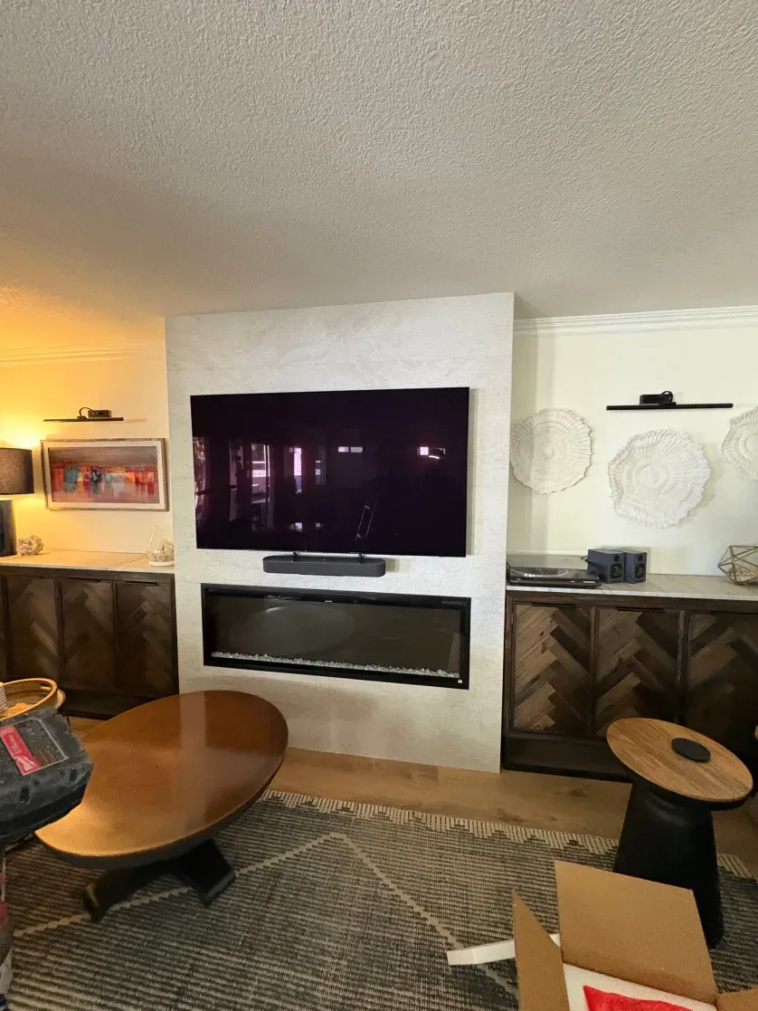 Living room with a mounted TV above a fireplace, flanked by cabinets. A coffee table and rug are in front.