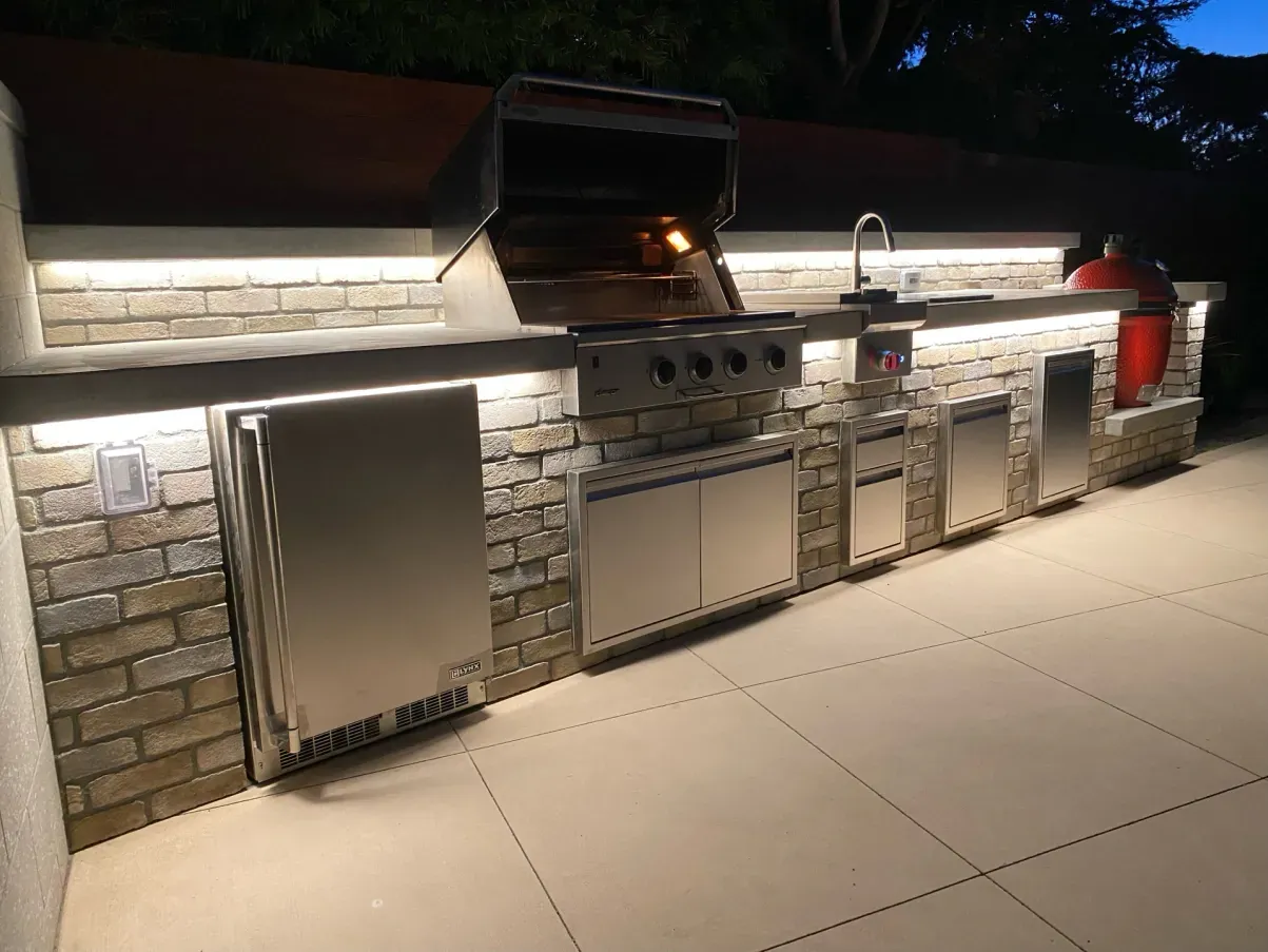 Outdoor kitchen with lit-up grill, sink, and storage, built into a brick-like structure, on a patio at dusk.