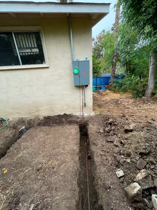Trench dug near a house with an electrical panel. Conduit runs from panel into the trench.