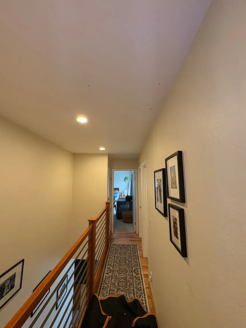 Hallway with beige walls, wooden railing, and a patterned rug. Framed artwork hangs on the wall.