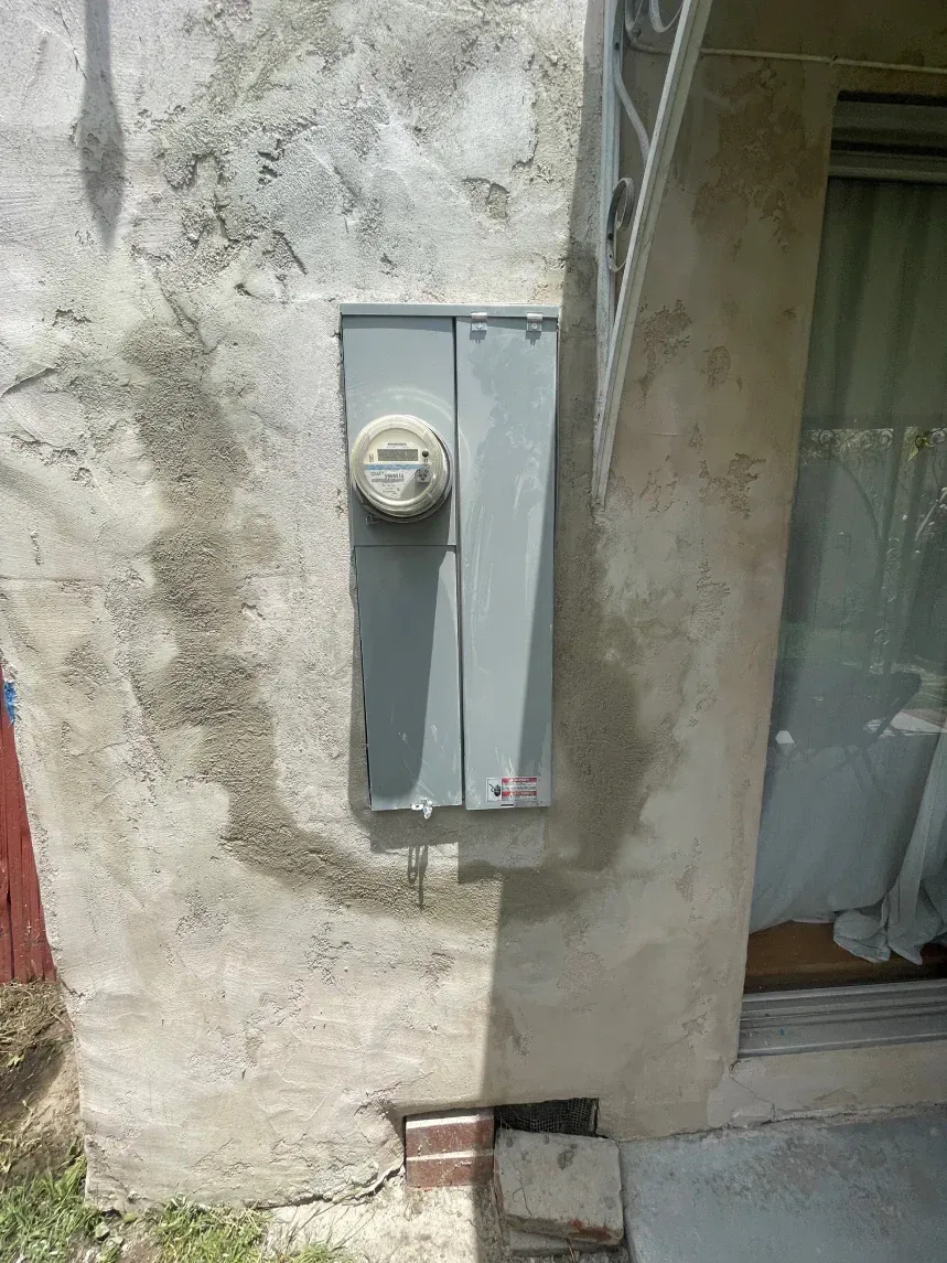 Electrical meter and panel on a stucco wall. Gray metal, dirt marks, brick footing, window to the right.