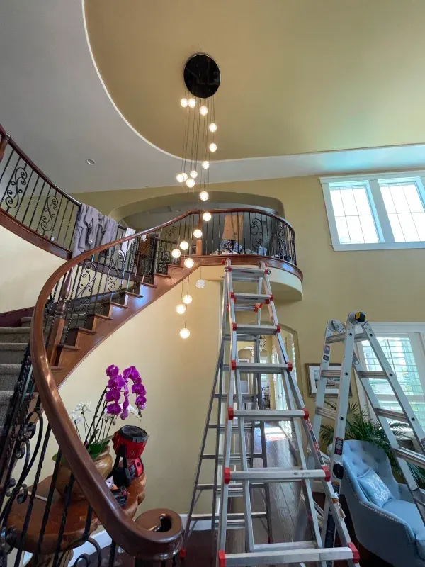 Spiral staircase with new chandelier installation, two ladders present, yellow walls.