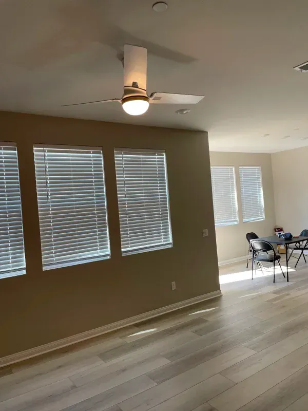 Living room with light wood floors, tan walls, windows with blinds, and ceiling fan.