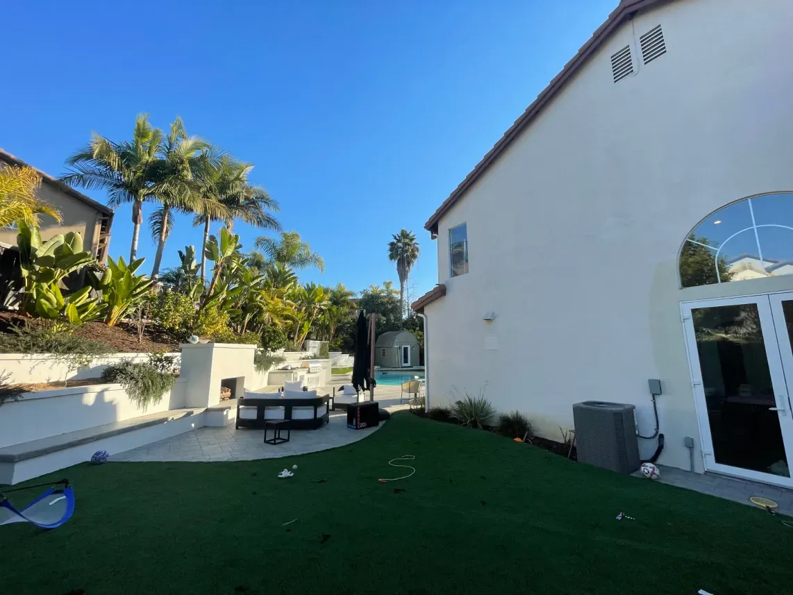 Backyard with artificial grass, patio furniture, pool, and a two-story beige house under a blue sky.