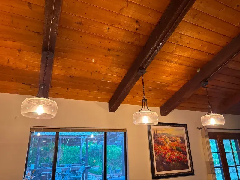 Three pendant lights hang from a wood ceiling with exposed beams. A painting and window are also visible.