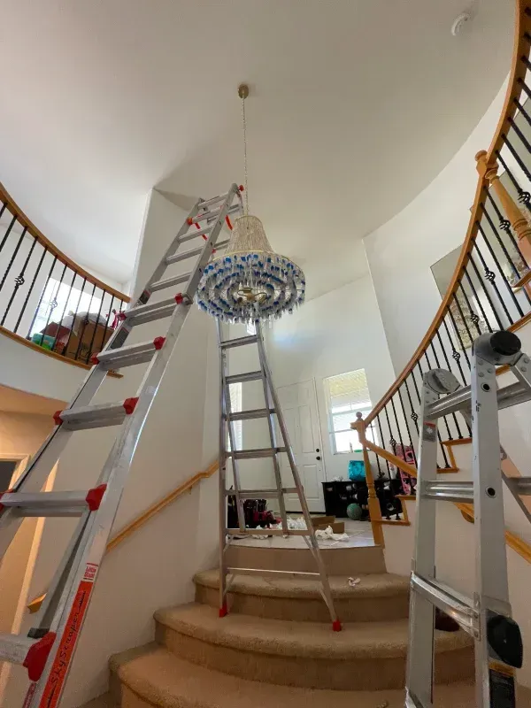 Three ladders set up near a staircase with a chandelier being worked on.