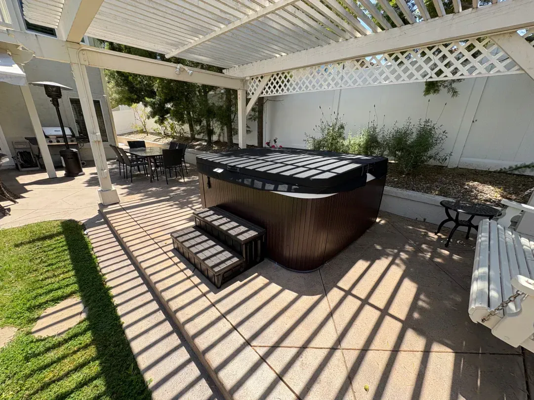 Hot tub under a white pergola on a concrete patio, with steps, and a white fence in the background.