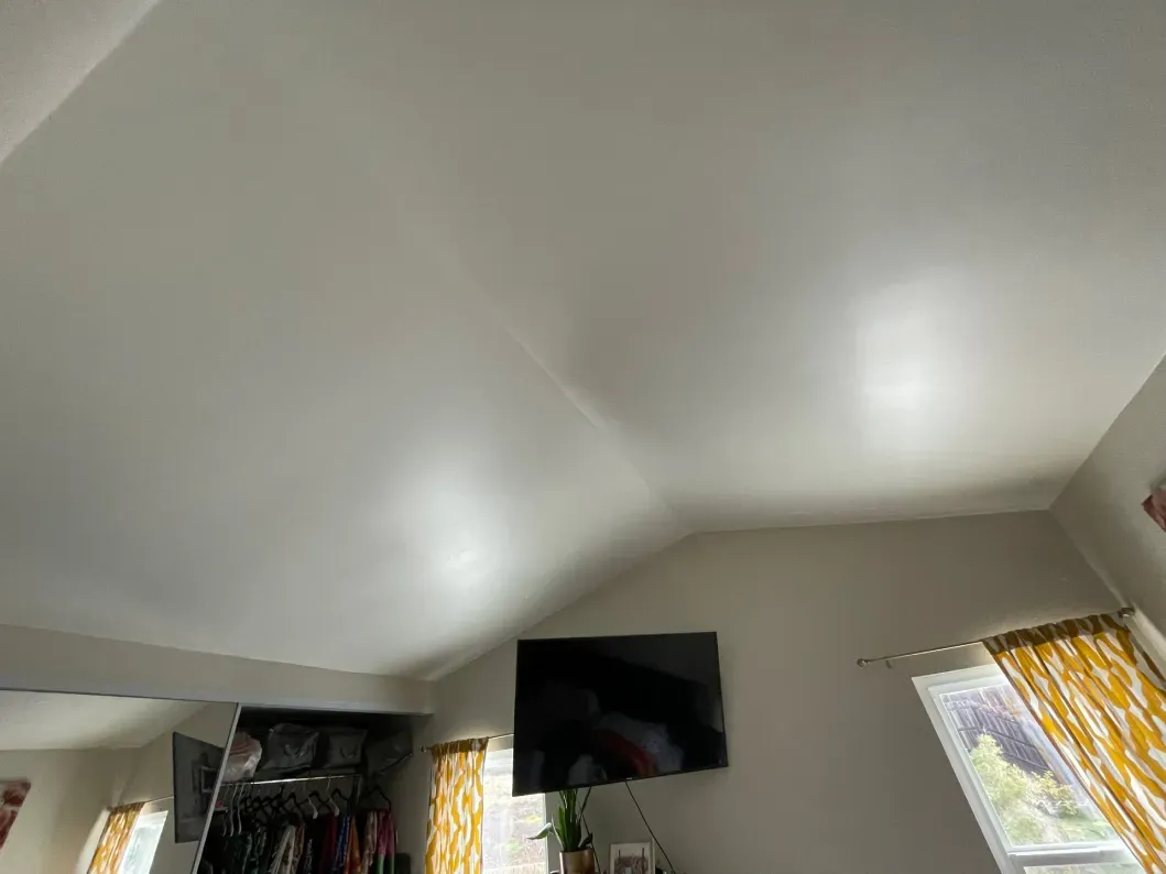 A room with a slanted white ceiling, a mounted TV, and a window with yellow curtains.