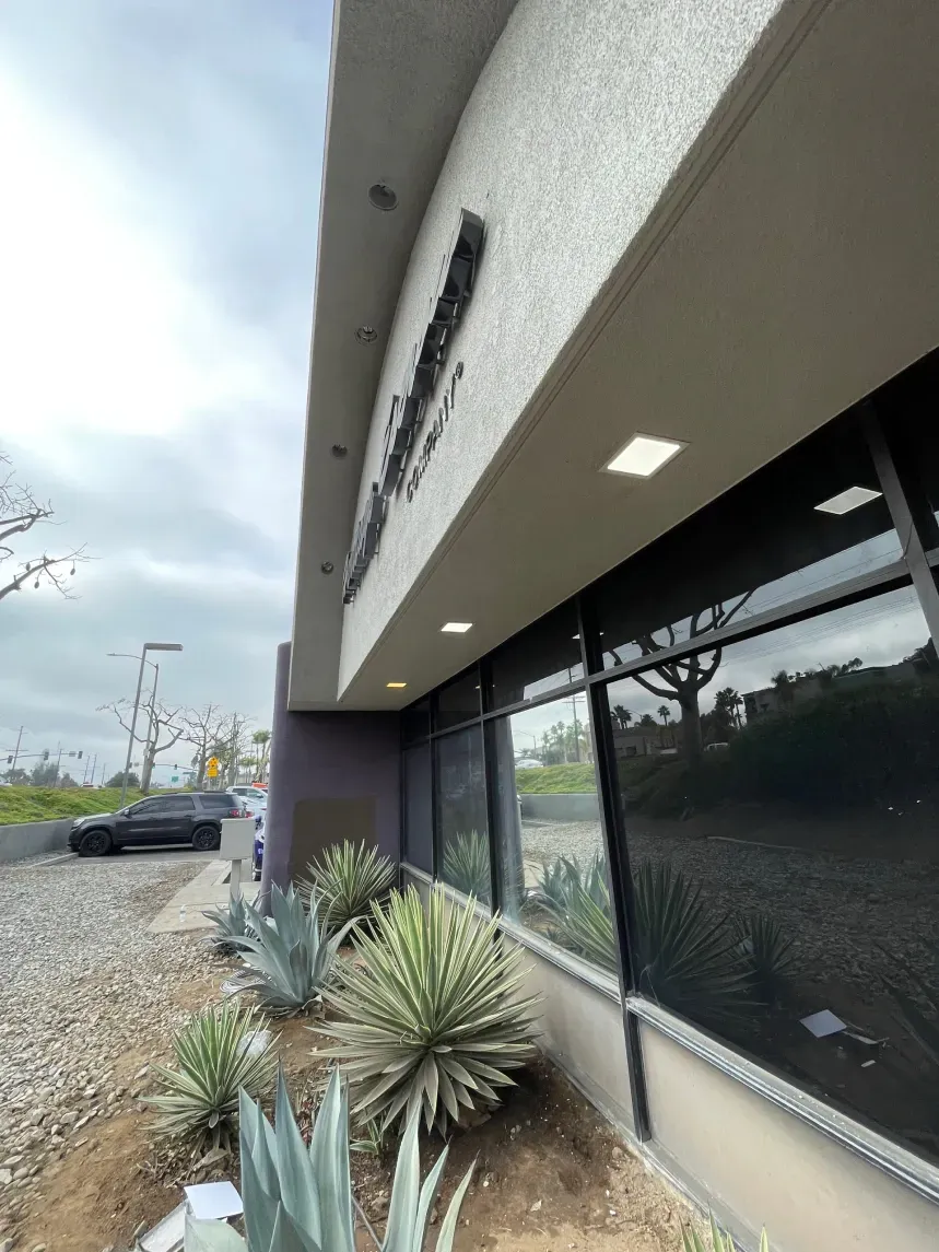 Exterior of a building with dark tinted windows, agave plants in front, and a cloudy sky.