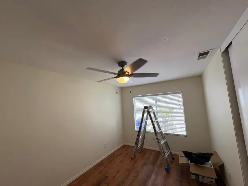 A room with a ceiling fan, ladder near a window with blinds, and hardwood floors.