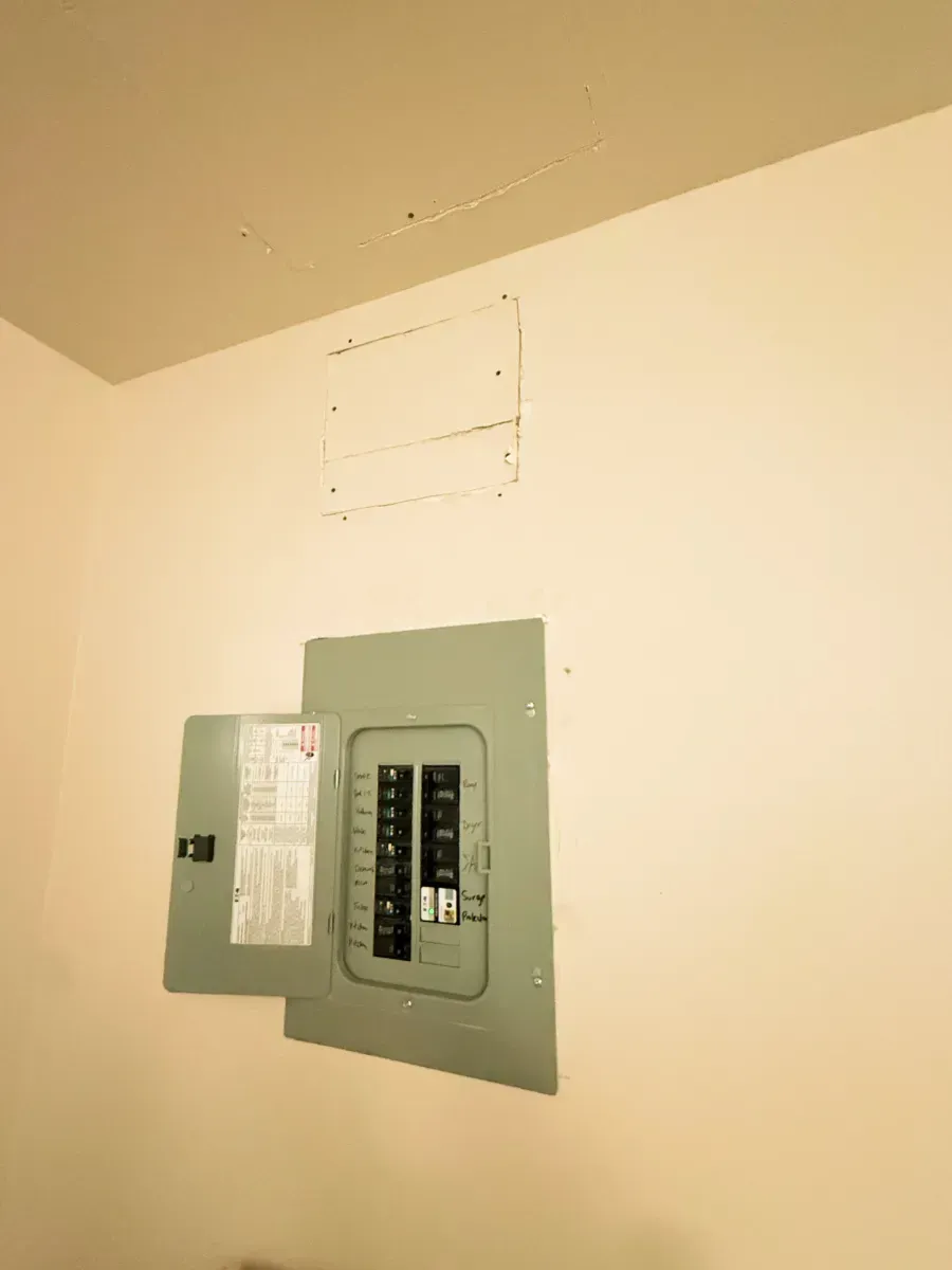 Electrical panel with open door on a white wall. The ceiling has a square cutout.