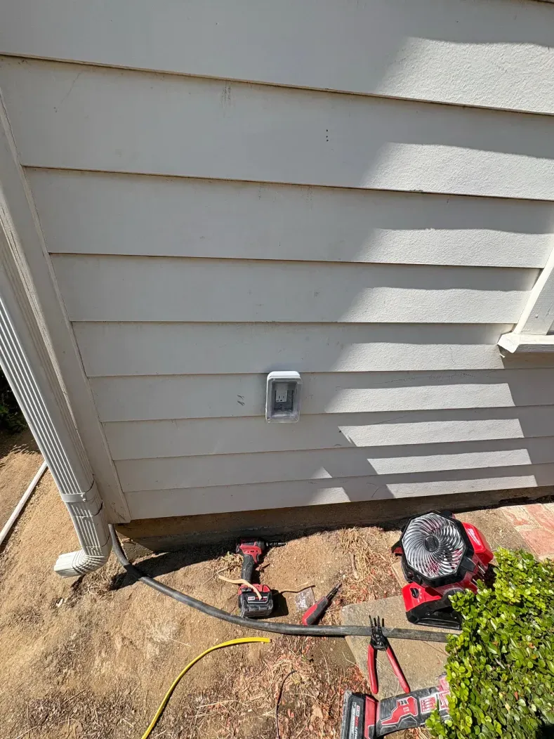 Exterior outlet on white siding, with tools and plants visible below. Sunny day.