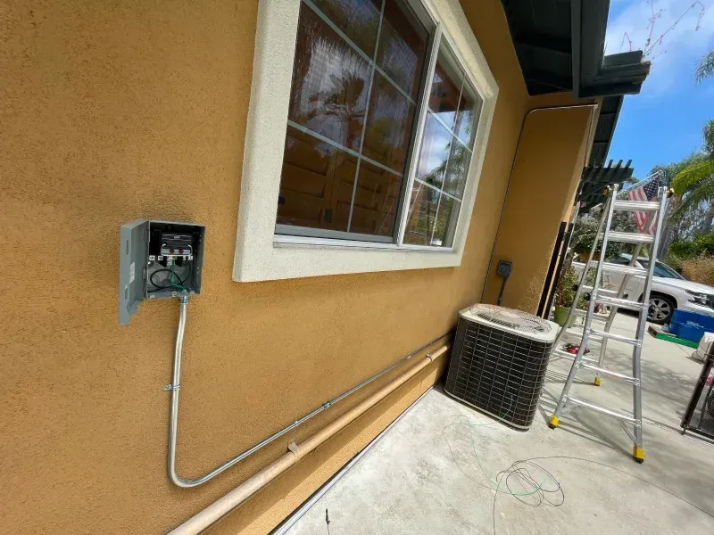 An electrical box and conduit on a stucco wall next to a window and air conditioner unit. A ladder is nearby.