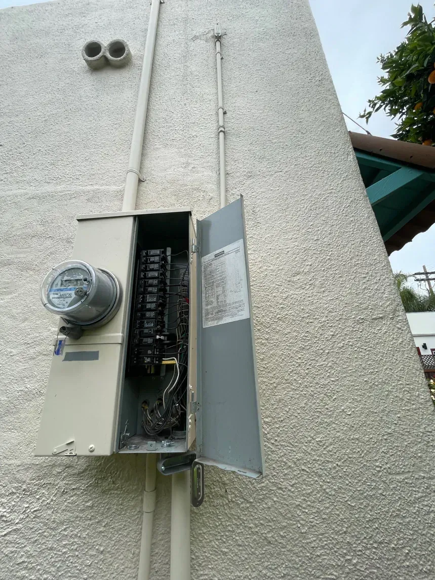 Exterior electrical panel with open door, showing fire damage and soot on stucco wall.