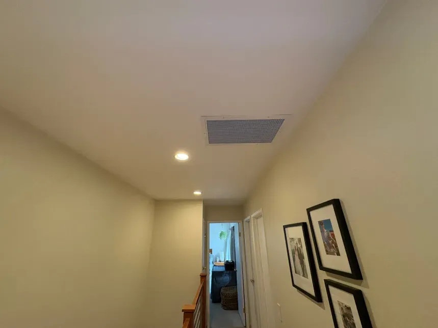 Hallway with cream-colored walls, recessed lights, and framed artwork. A vent is in the ceiling.