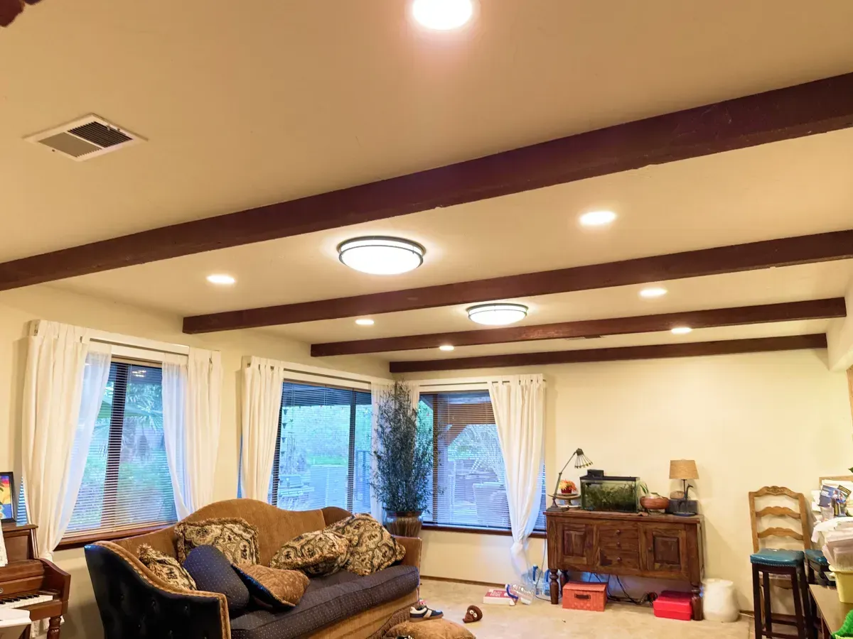 Living room with cream-colored walls, dark wooden beams, and recessed lighting. Windows with white curtains and a dark couch.