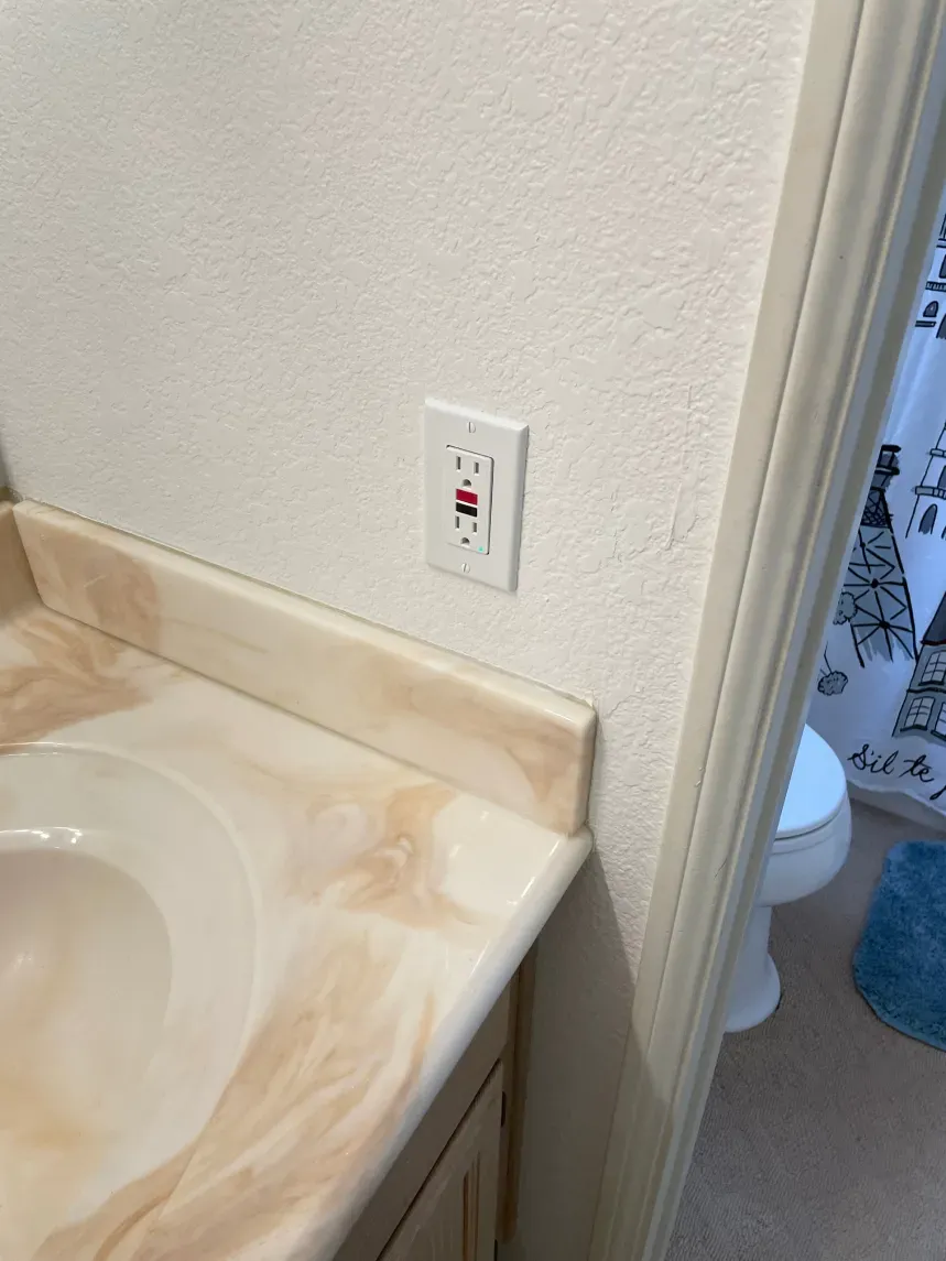 White electrical outlet with a red button on a bathroom wall next to a sink.