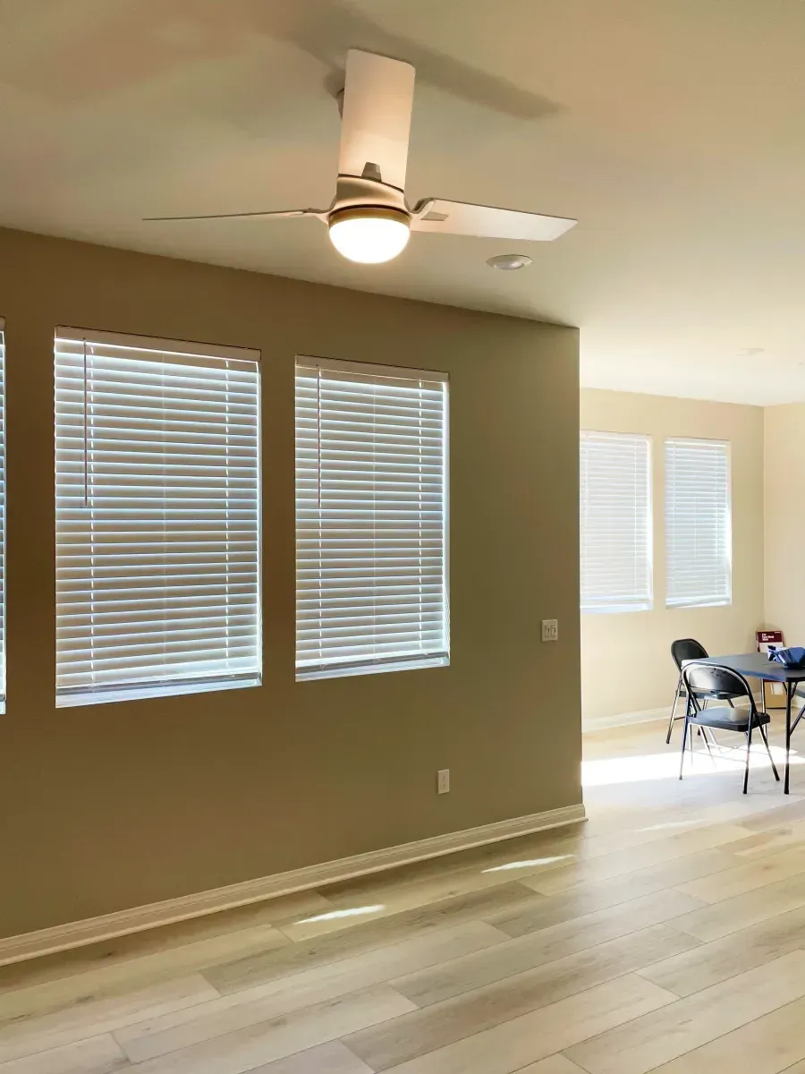 A room with a ceiling fan, three windows with blinds, and light wooden flooring.