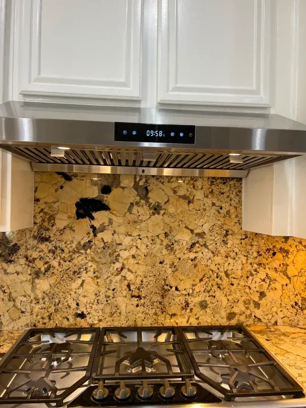 Stainless steel range hood above a gas cooktop and granite backsplash with white cabinets.