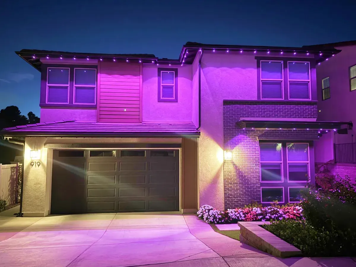 Two-story house illuminated in purple. Garage door closed, windows visible. String lights across the roof.