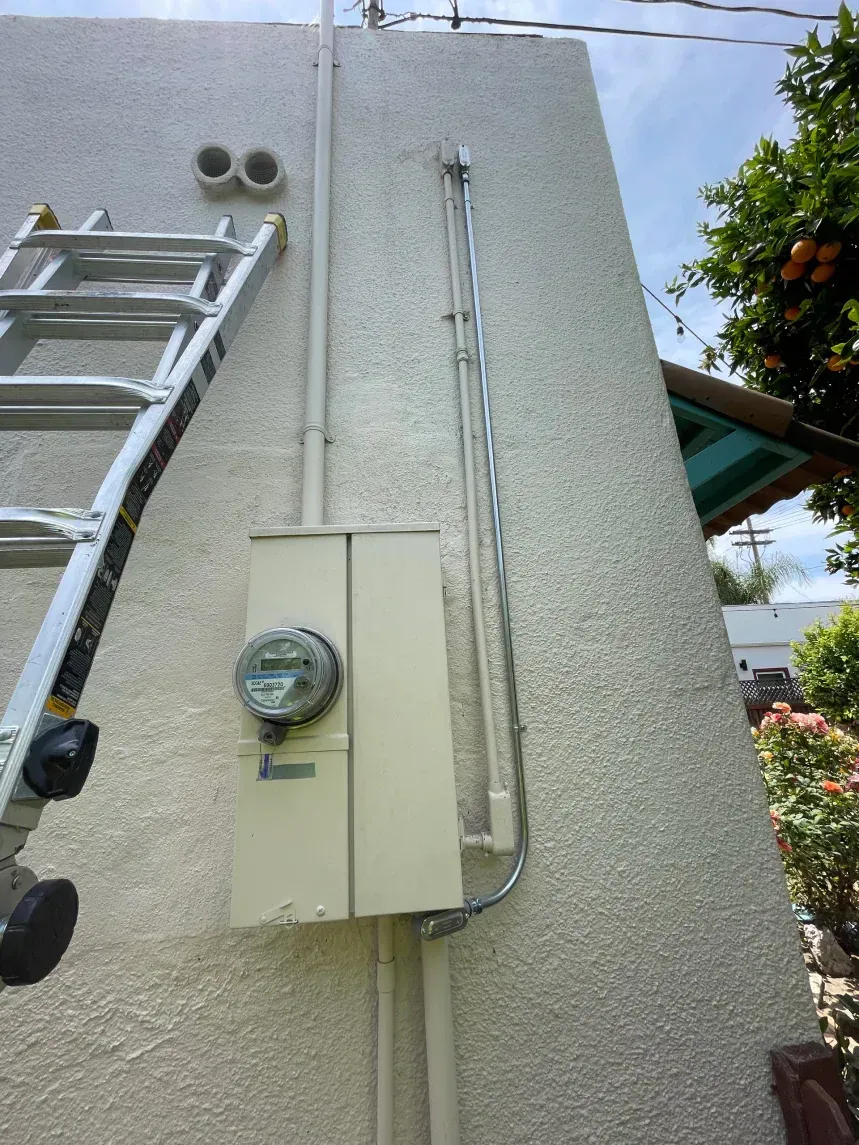Ladder next to a stucco wall with electrical meter box, pipes, and utility lines. Sunny outdoors.
