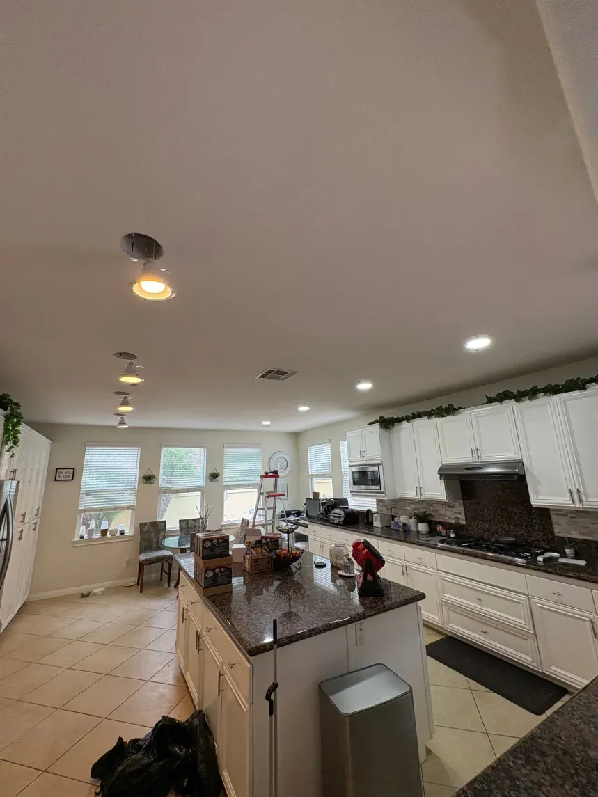 Bright kitchen with white cabinets, island, and ceiling lights; windows and appliances visible.