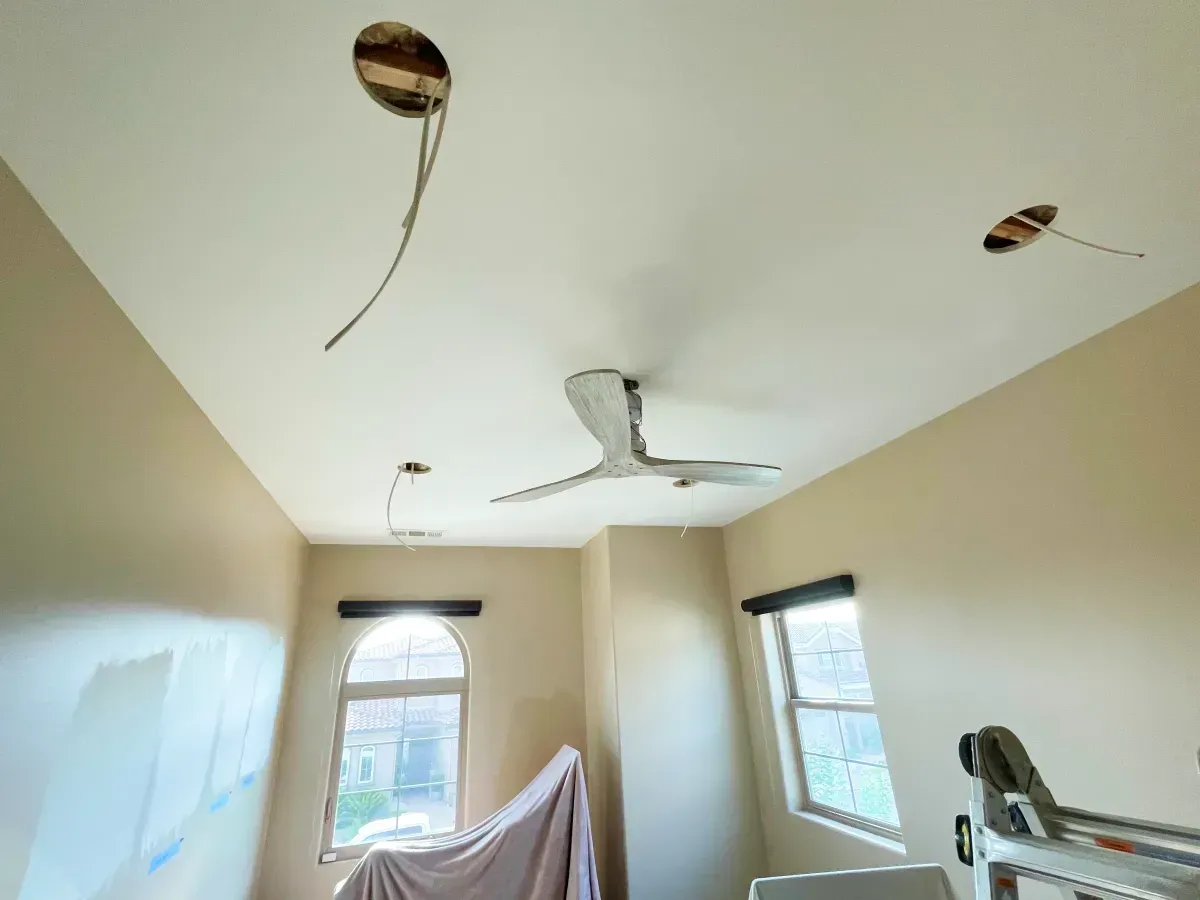 Room with a ceiling fan, exposed wiring, and holes. Windows, a tarp-covered object, and a ladder are visible.
