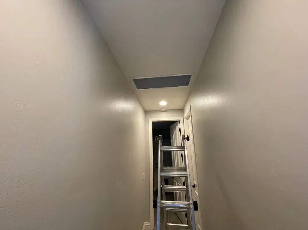 Narrow hallway with a ladder, an open ceiling panel, and a doorway. Beige walls and a white ceiling.