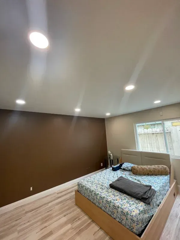 Bedroom with brown accent wall, bed, and recessed ceiling lights. Light wood flooring and a window.