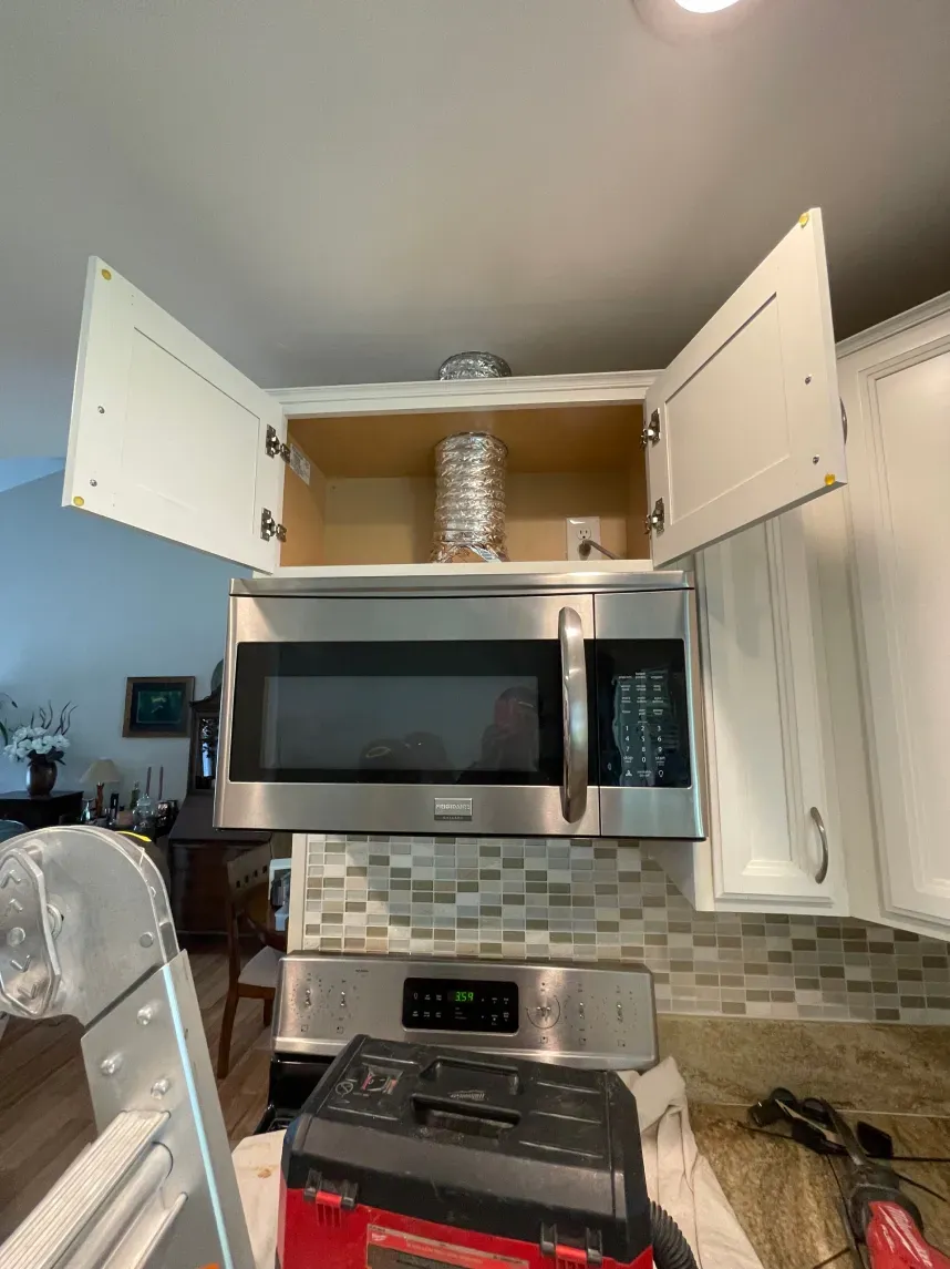 Microwave installed under a cabinet in a kitchen with open doors, showing vent and tools below.
