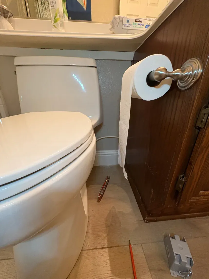 Toilet, toilet paper roll, and wooden cabinet in a bathroom. Pencil and electrical component on the floor.